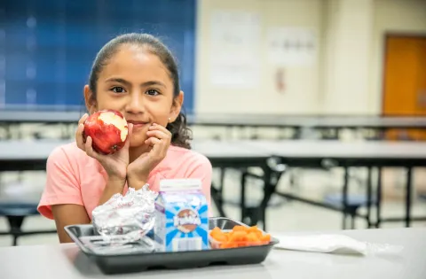 Young Latina child in a school cafeteria holding an apple.