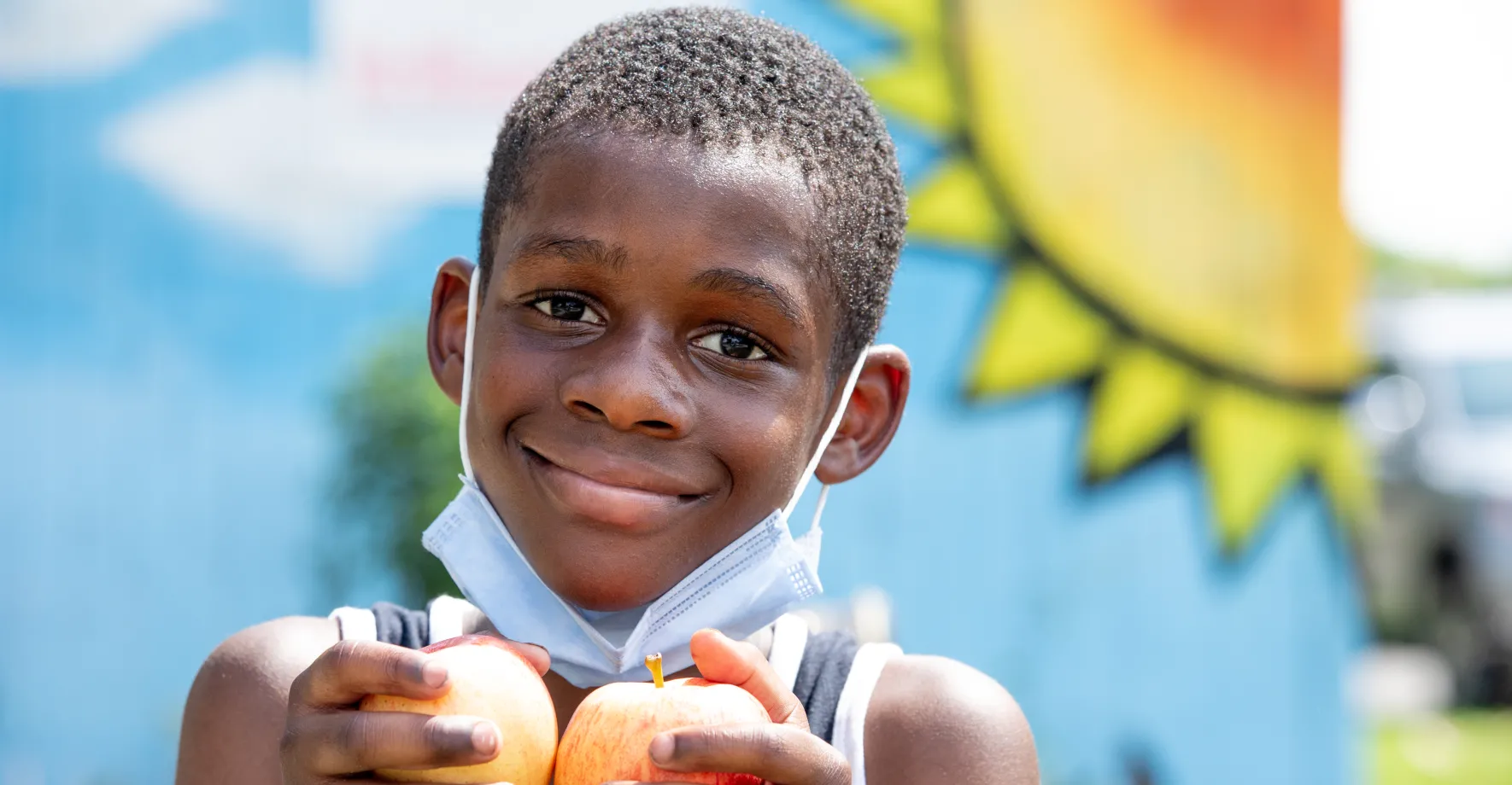 Black kid with mask under the chin holding two apples and happy