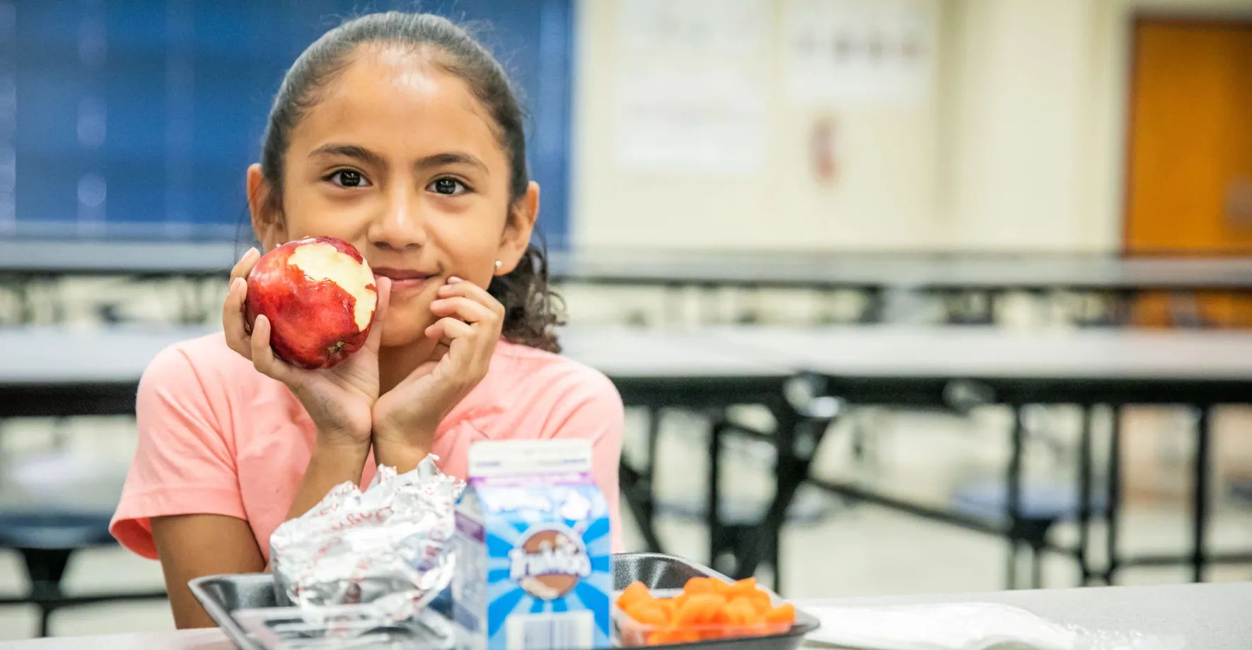 Young Latina child in a school cafeteria holding an apple.