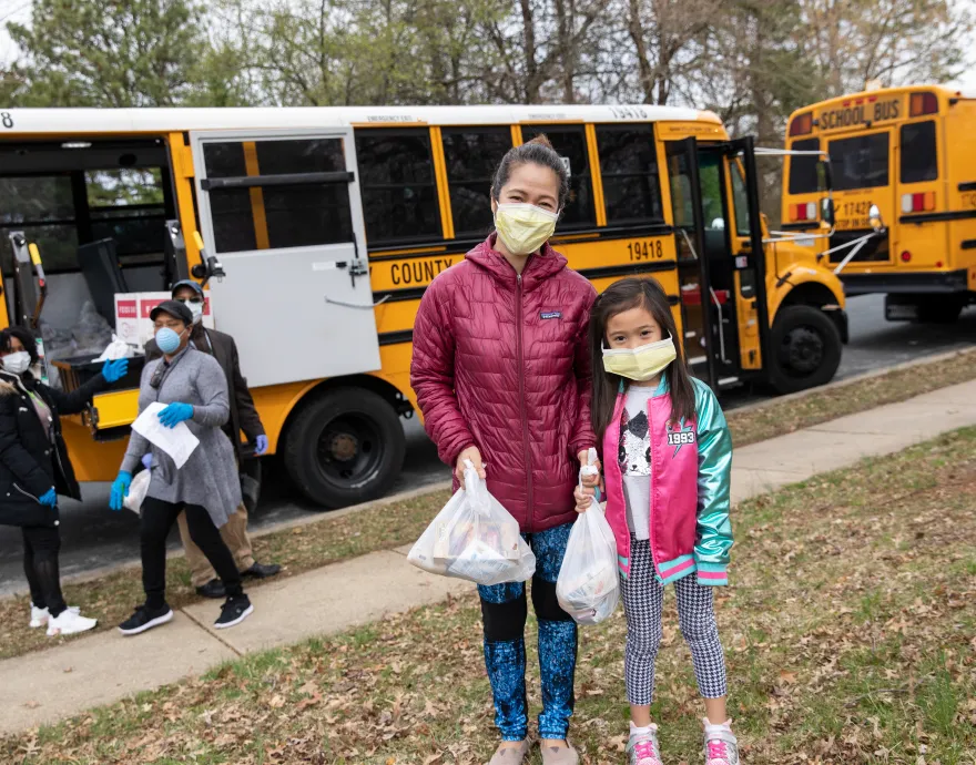 A mother and daughter wear face masks and carry their meals away from the school bus.