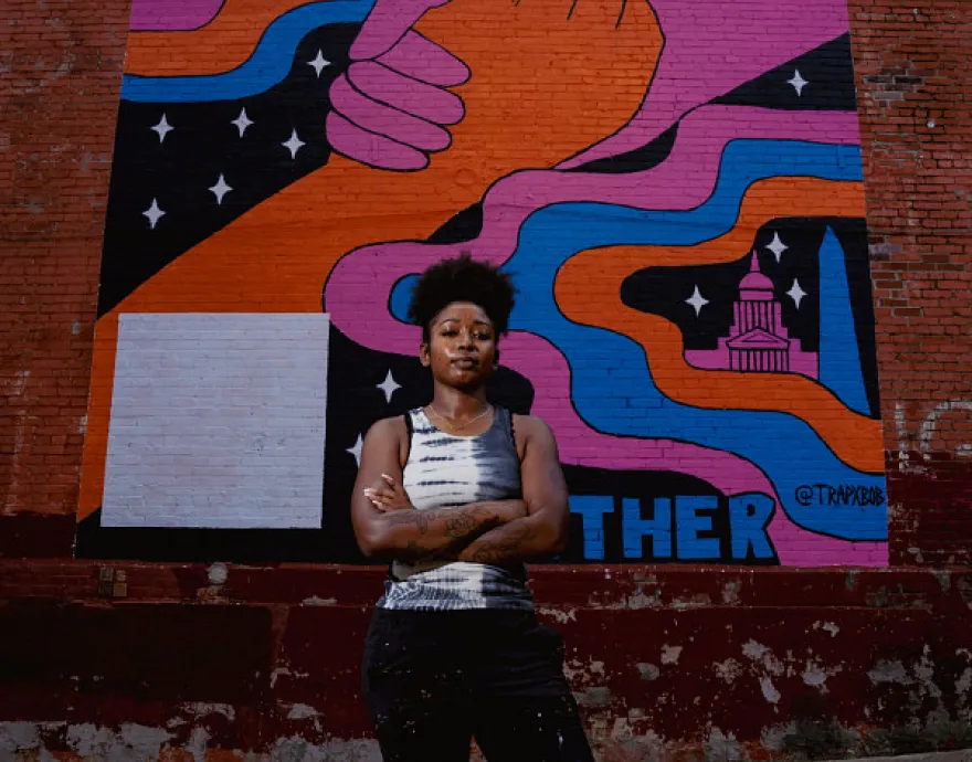 Artist Trap Bob stands in front of her mural with her arms folded and covered in specs of paint.