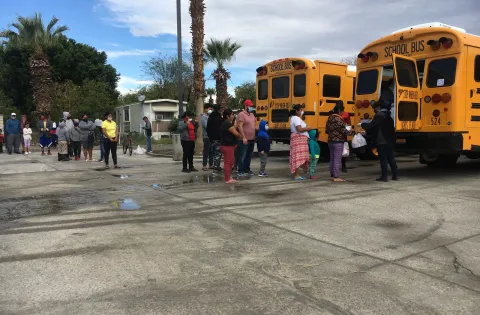 A line of people wait for food in a parking lot behind two school buses.