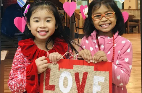 Two girls holding a sign that says love is all you need. They have hearts in the background
