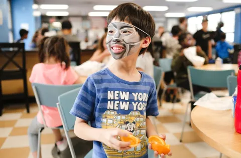 Smiling boy with face paint in crowded cafeteria holding orange slices
